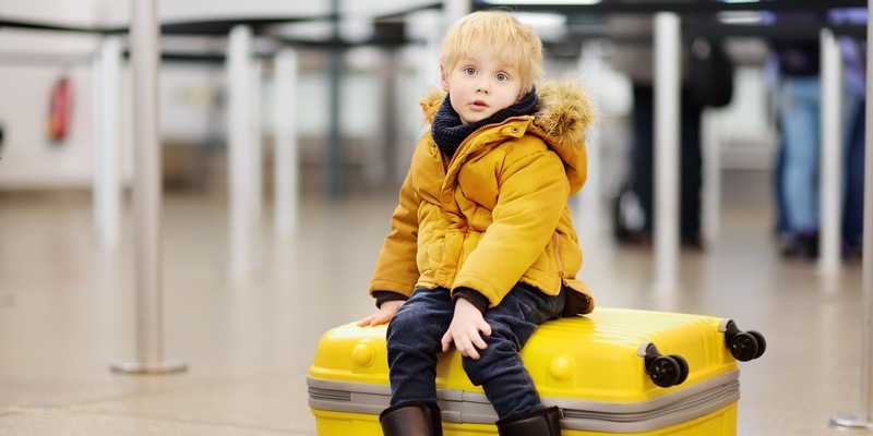 kleiner Junge sitzt wartend am Flughafen auf einem gelben Koffer