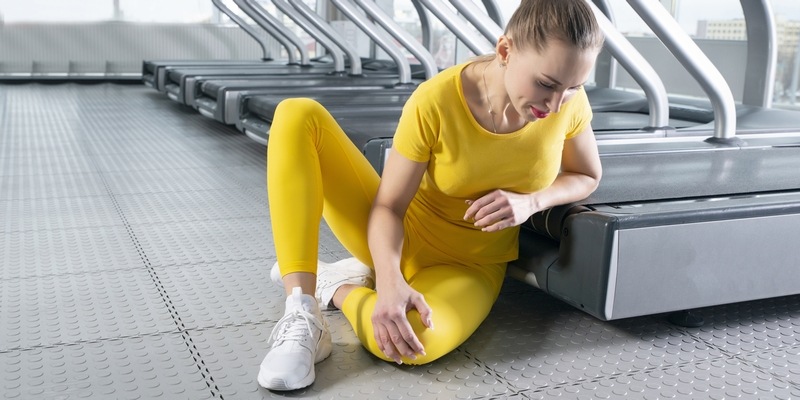 junge Frau vor Laufbändern hält sich verletztes Knie